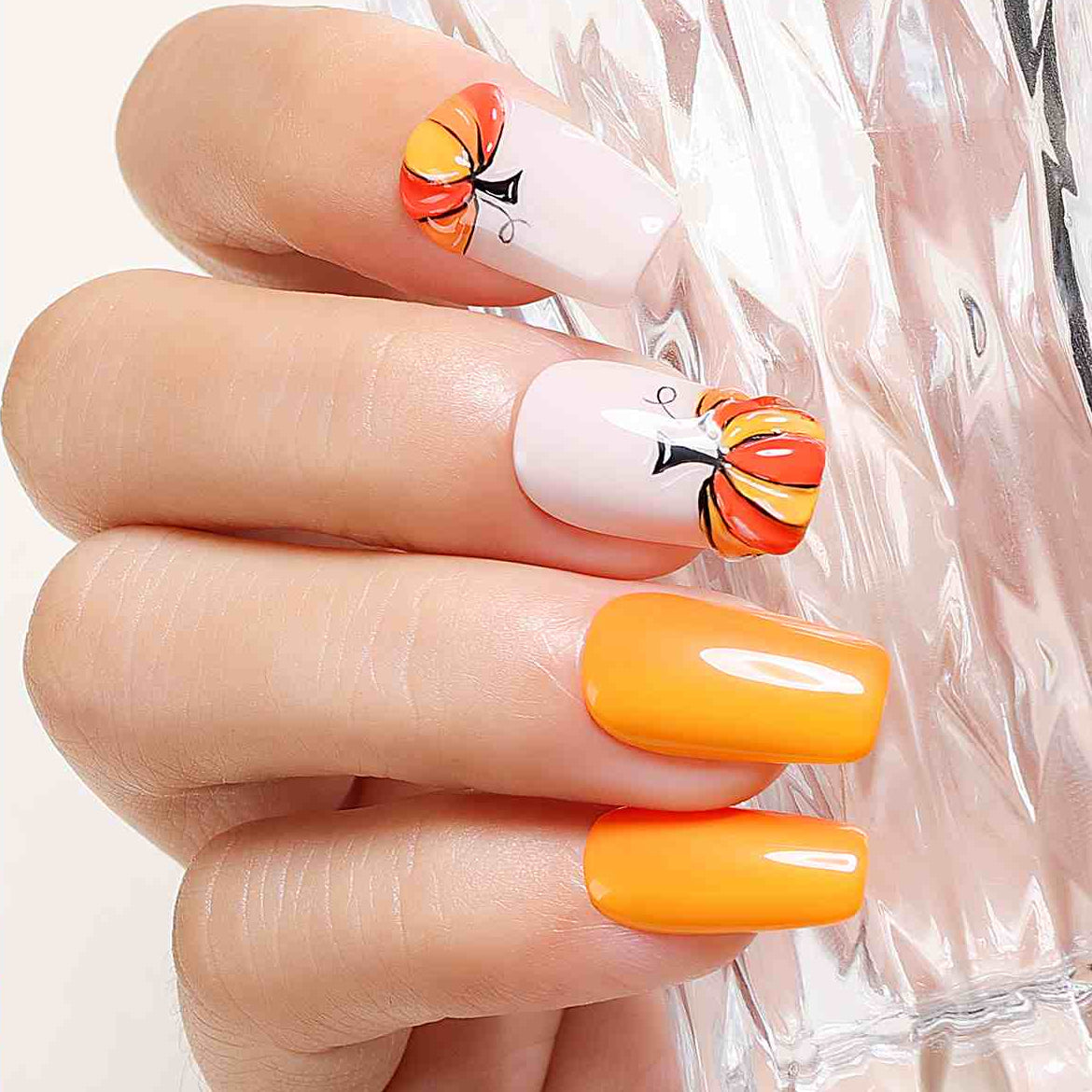 A close-up of a hand showing manicured nails with a glossy orange-yellow color and two white nails decorated with small orange and yellow pumpkin designs. The image includes promotional text about creating custom nail styles and mentions multiple color options.